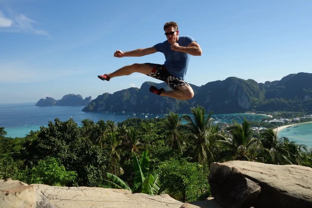 Rick Tew in a flying kick at the Koh Phi Phi viewpoint, green limestone islands and turquoise bays behind him
