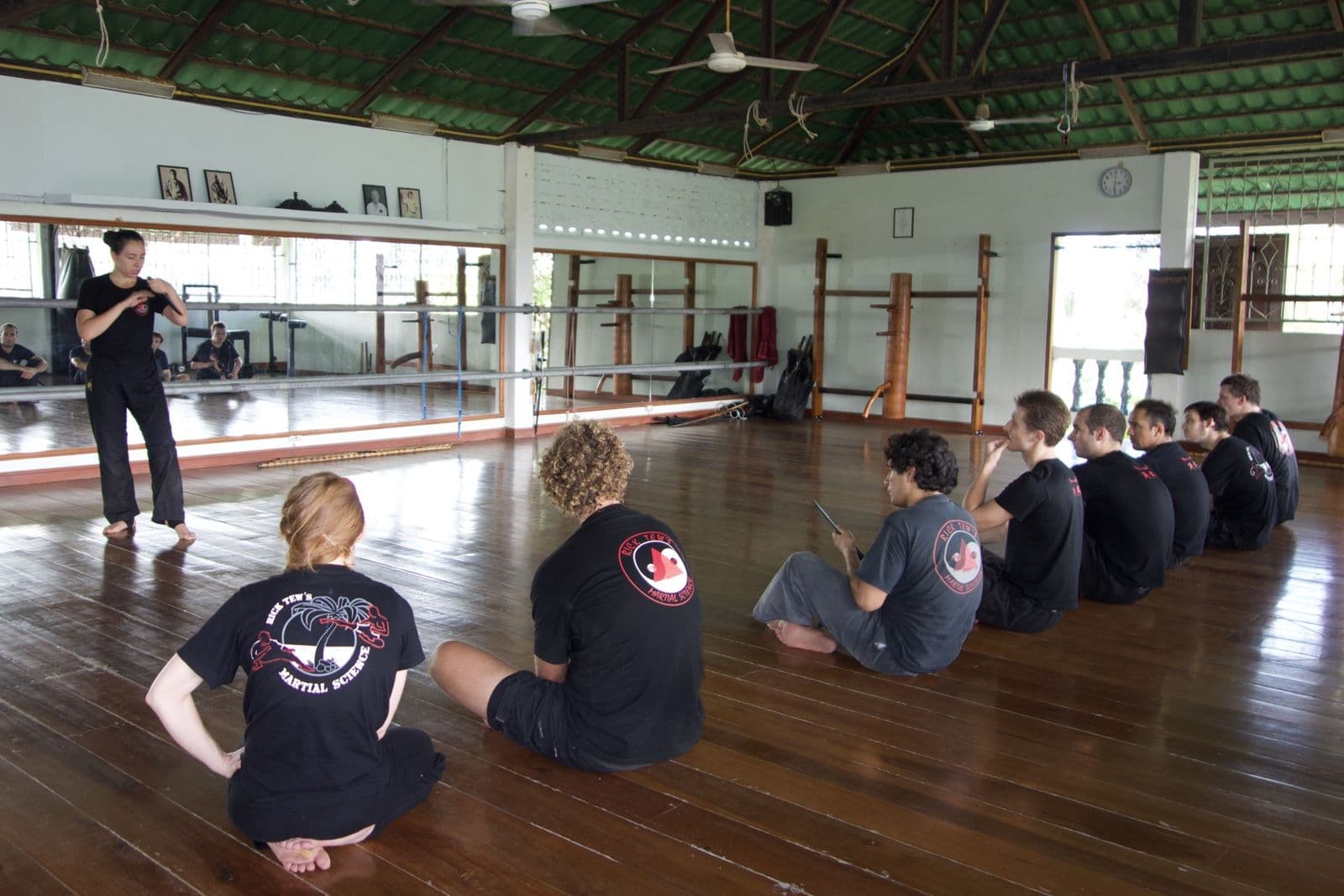 A row of students sitting on a wooden floor watching a woman instructor demonstrate