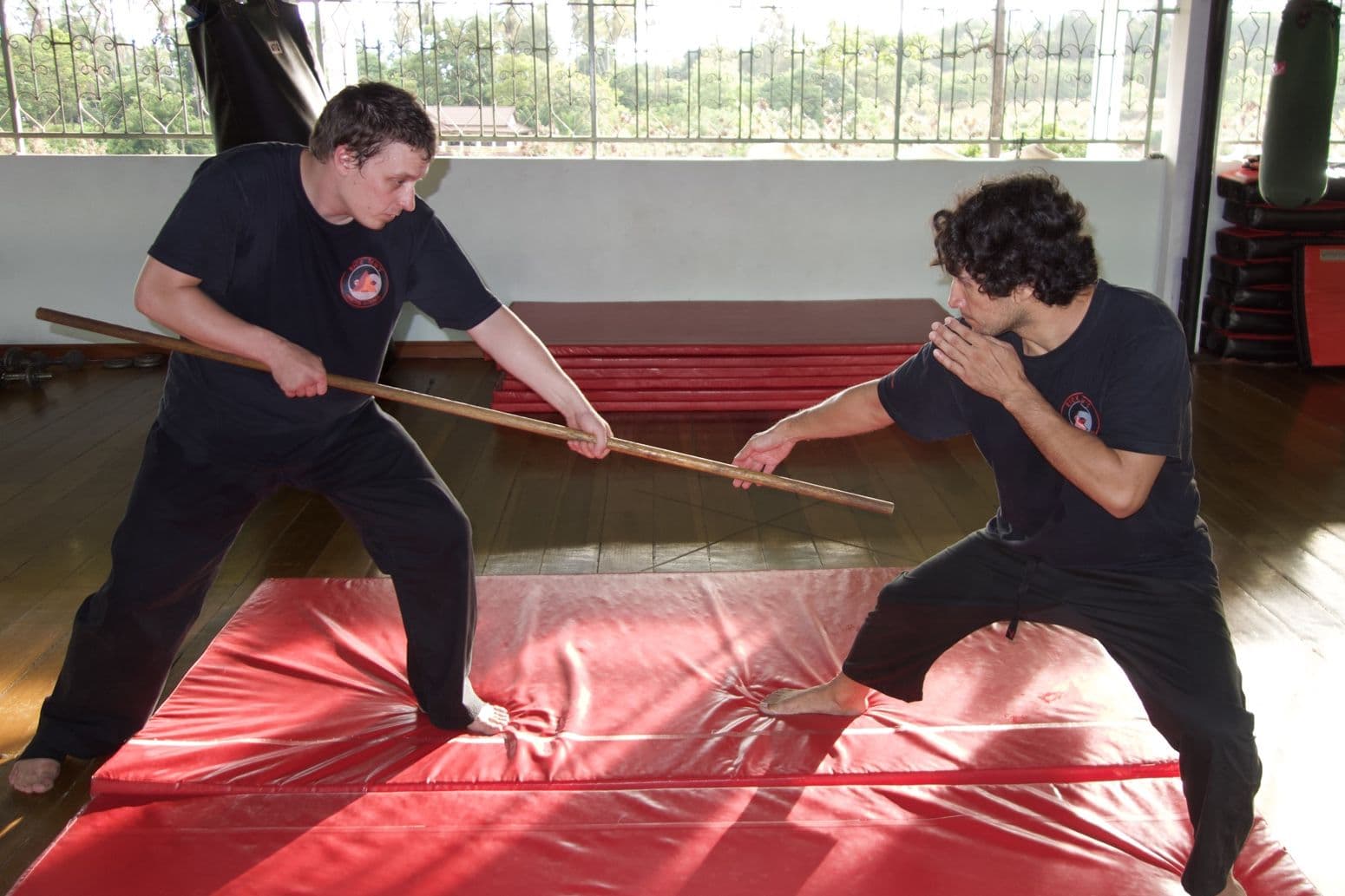 Two students in low stances crossing a long staff on red mats in an open-sided wooden hall