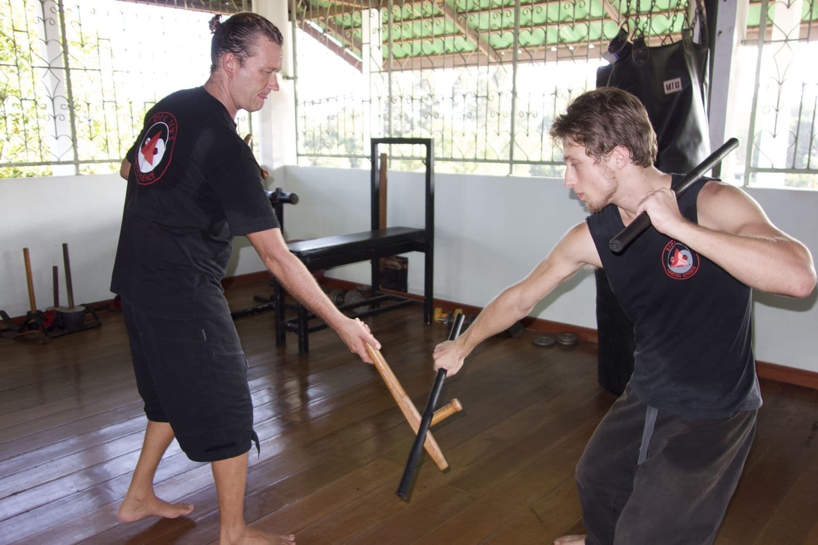 An instructor and a student practicing with short sticks on a wooden floor, trees through the windows
