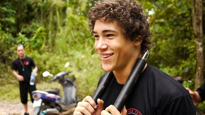 A smiling student holds his nunchaku on a jungle trail, scooters parked behind him
