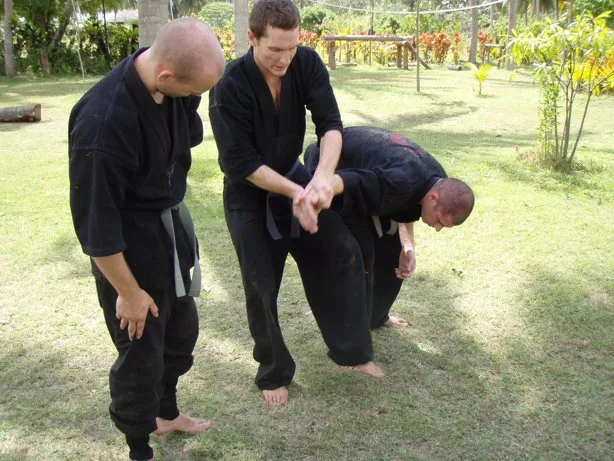 On a garden lawn, a student helps two classmates through a wrist hold during review