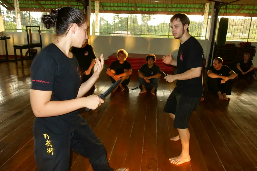 A woman works a stick drill with her partner while the class watches