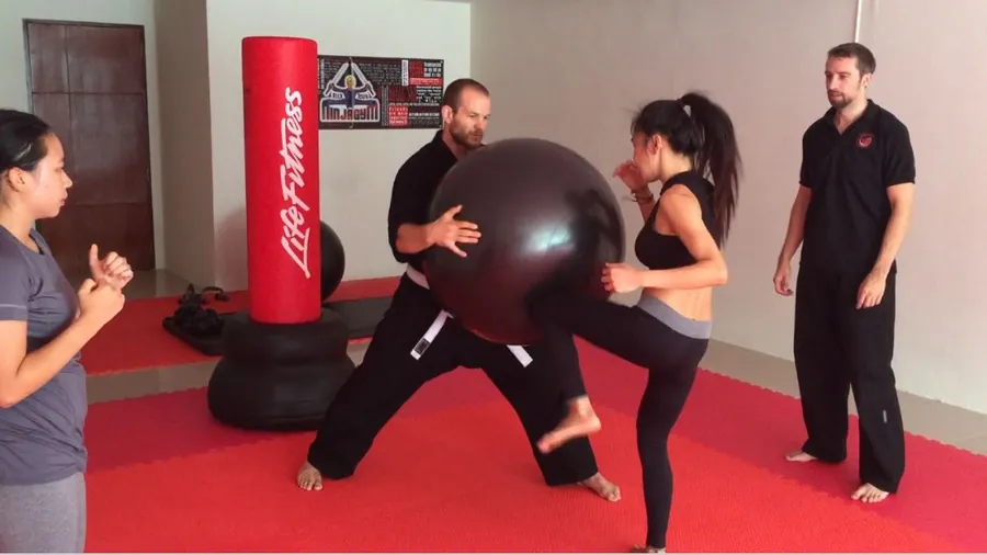 A woman drives a knee into a big exercise ball her partner holds