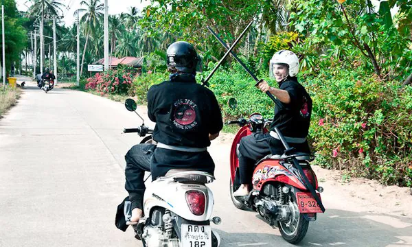 Two students in helmets ride scooters down an island road