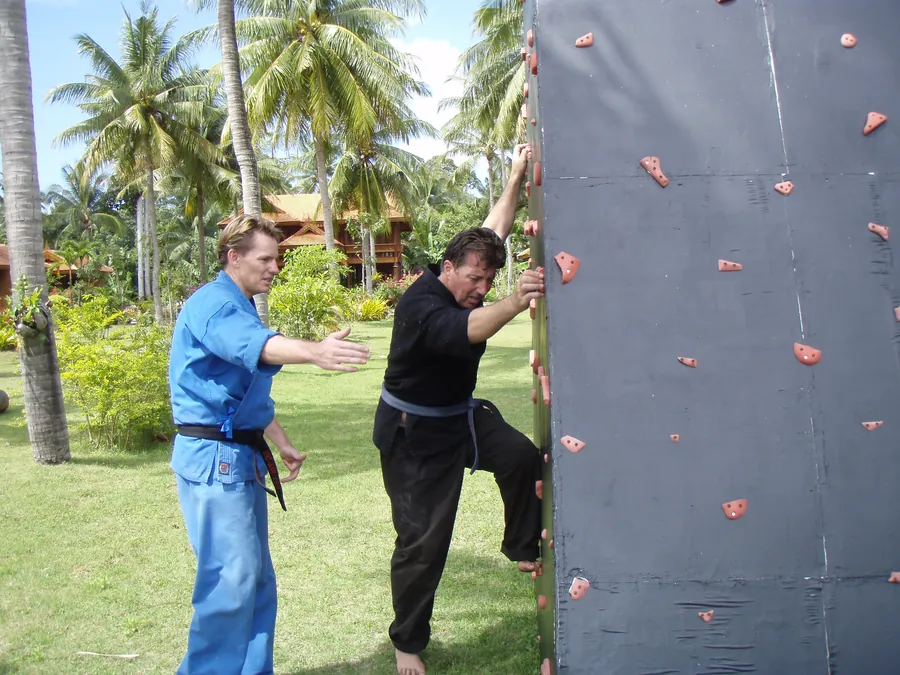 Rick Tew in a blue uniform coaches a student at a climbing wall in a garden
