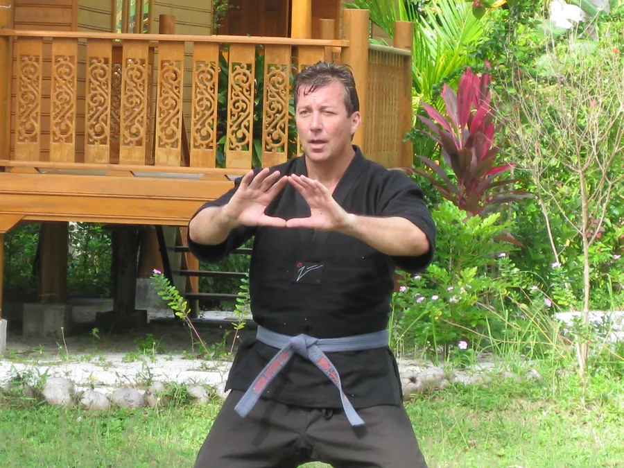 Rick Tew in a black uniform shows a hand position in a garden by a teak house