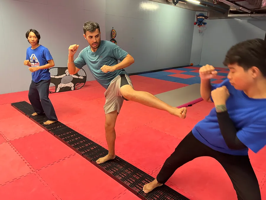 A student throws a round kick in a bright gym with red and blue mats