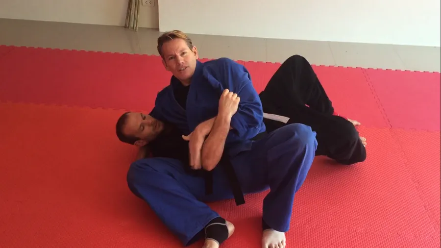 Rick Tew in a blue uniform pins a student with an arm hold on red mats
