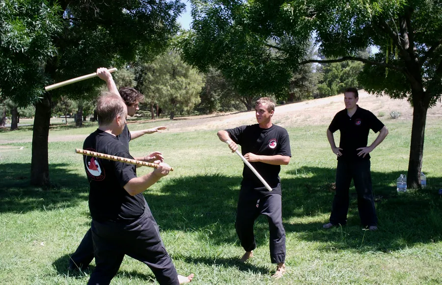 Students in black swing sticks in a park while their partners block