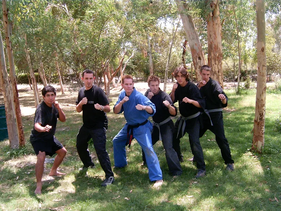 Rick Tew in a blue uniform and five students in fighting stance among eucalyptus trees