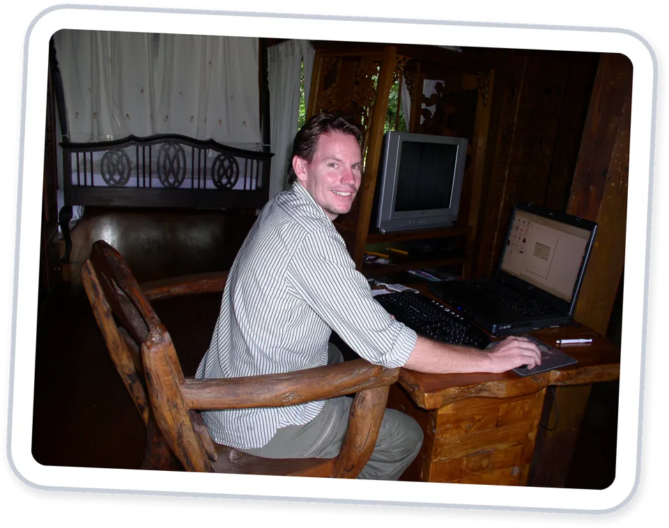Rick Tew smiling at a wooden desk with a laptop, in a treehouse room in Pai